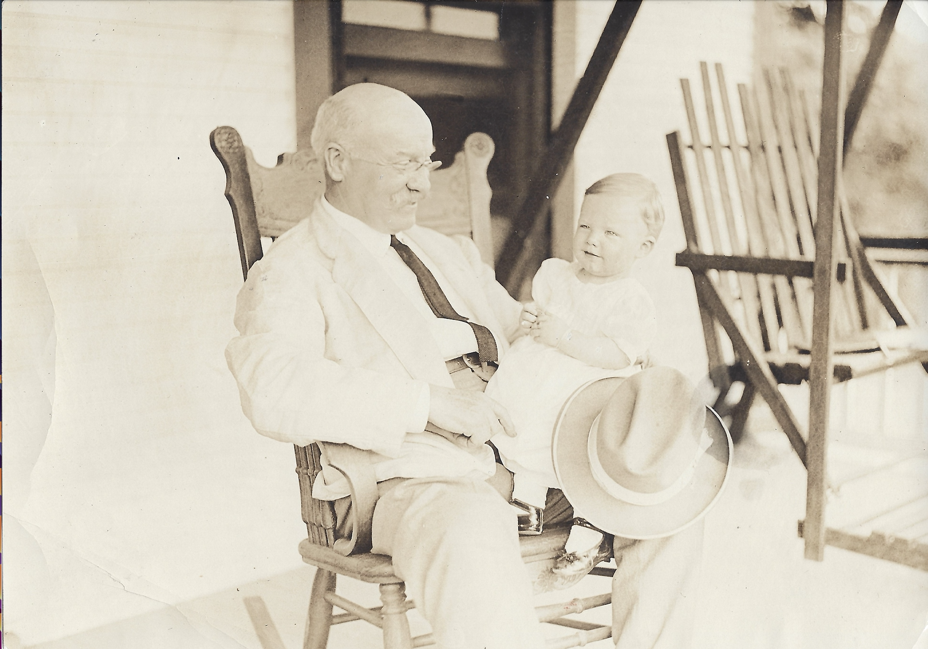 Frank Wyman sits in a rocking chair on a porch. A small child sits on his lap.