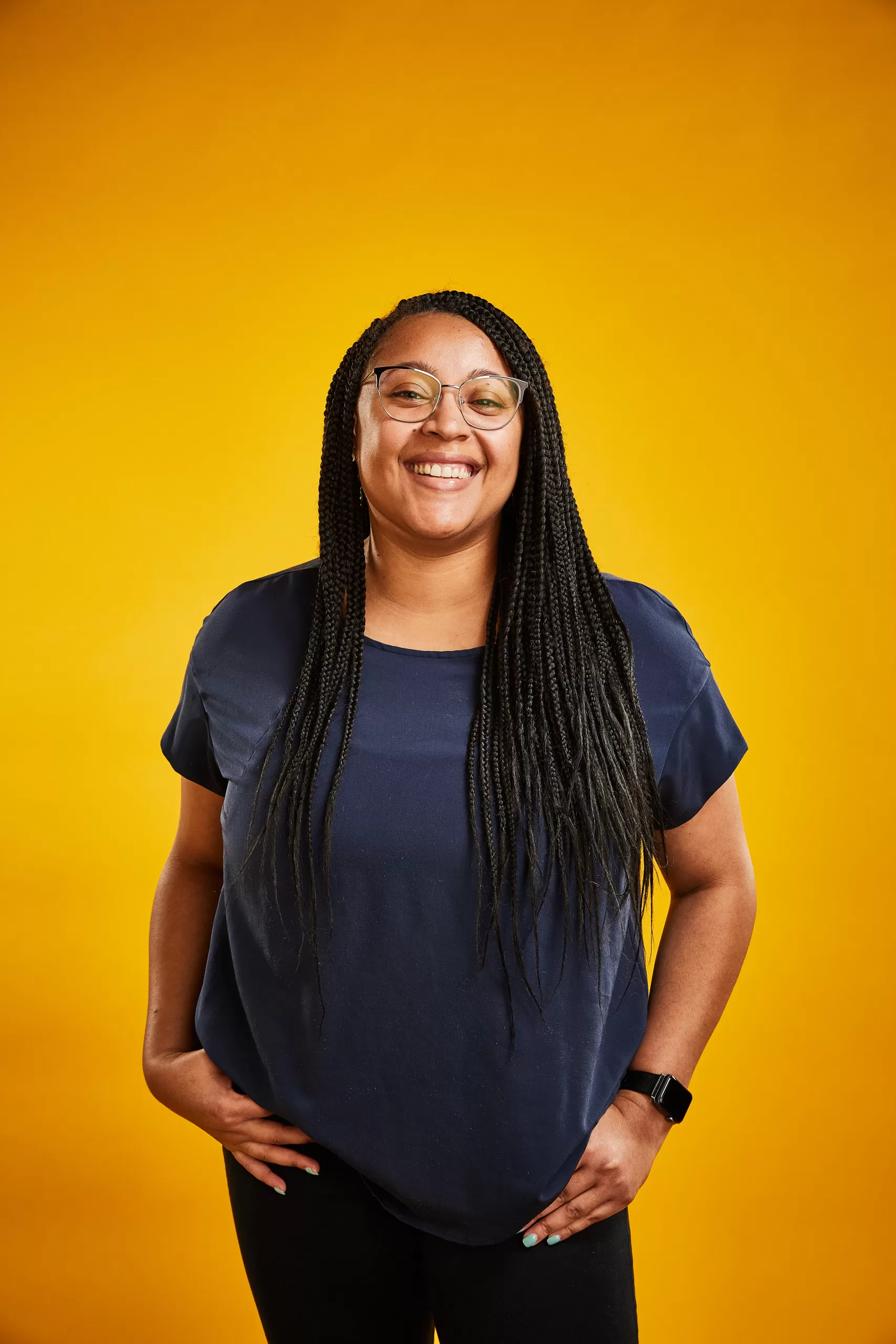 A young woman with braided hair, wearing a blue shirt, smiles to the camera in front of a yellow background.