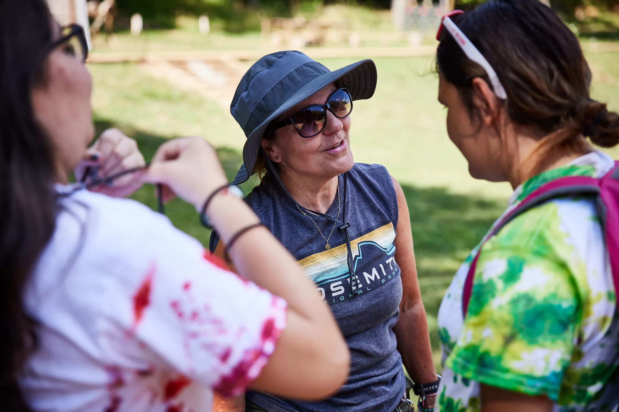 An outside scene where a female camp counselor wearing a hat chats with two campers wearing tie-dye shirts.