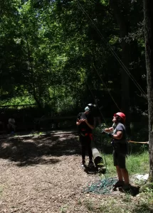 A counselor belays a climber on a rope while another camper watches next to her.