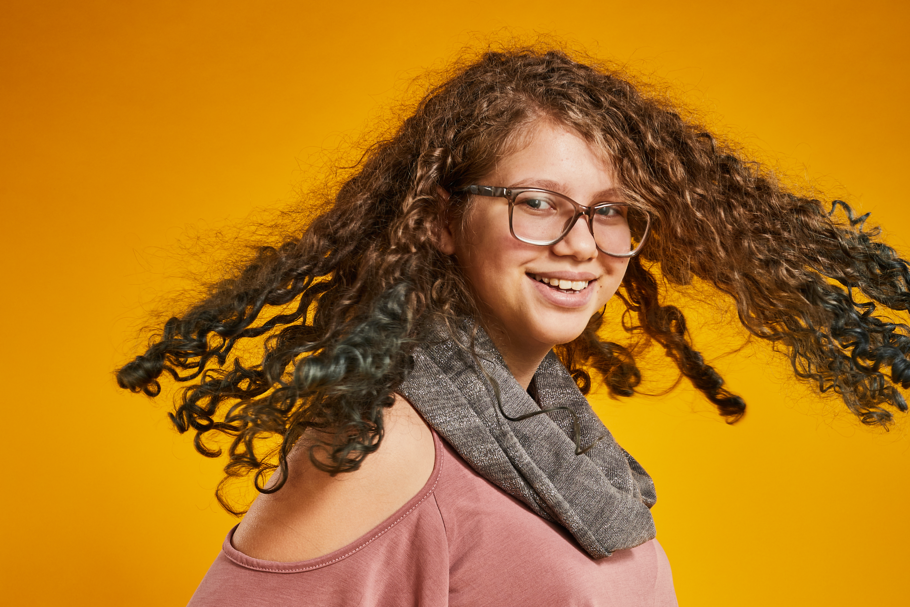 a young woman in front of a yellow background smiling at the camera as she swings her hair out