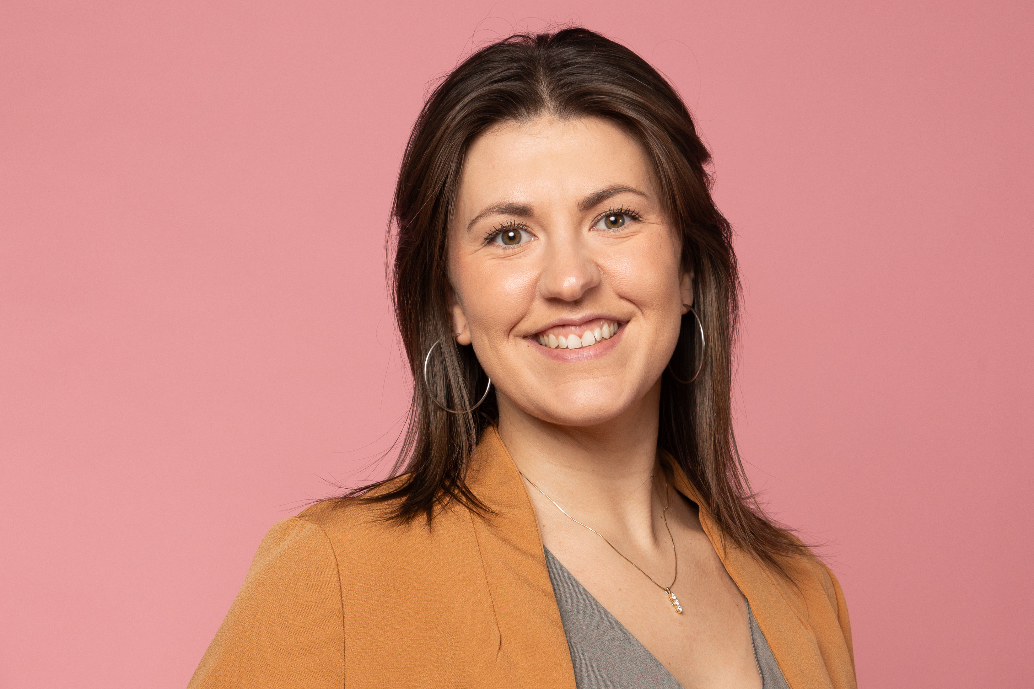 a young woman poses in front of a pink background smiling at the camera