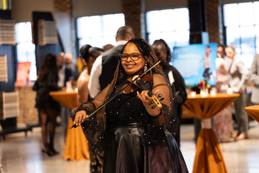 a woman with long braids and sparkly, sheer black dress stands at an event playing the violin