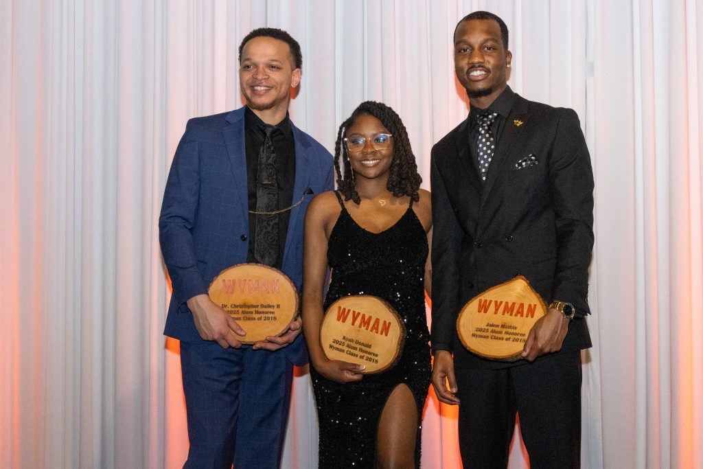 Two young men stand beside a young woman, all dressed in fancy attire and holding wooden plaques honoring them for their achievements
