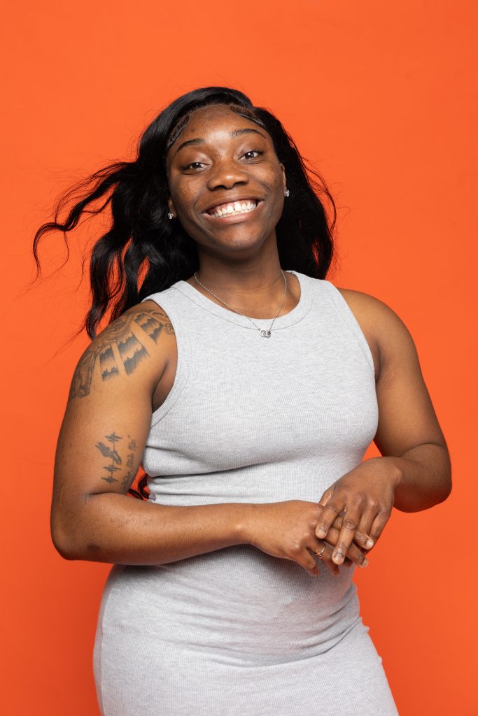 A young woman in front of an orange backgroud poses smiling at the camera