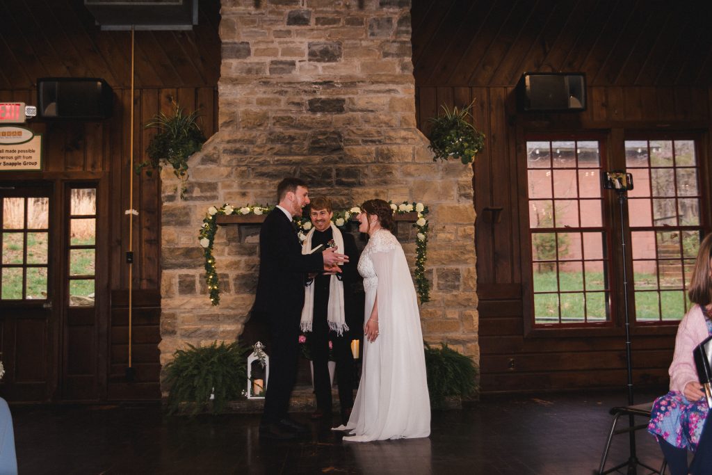 A wedding couple and their officiant stand indoors in front of a stone fireplace, smiling at one another.