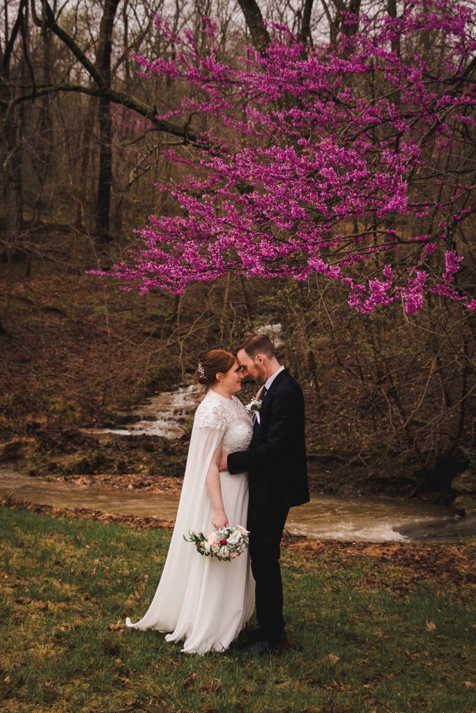 A wedding couple stands in an embrace under a tree with red-purple leaves