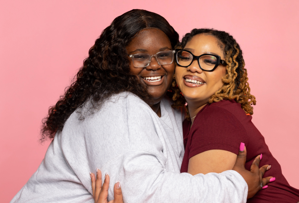 two young women hug and smile at the camera in front of a pink background