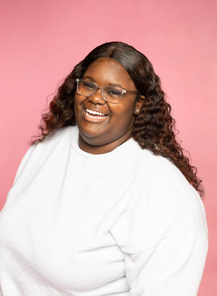 a young woman wearing glasses and a white shirt smiles at the camera in front of a pink background
